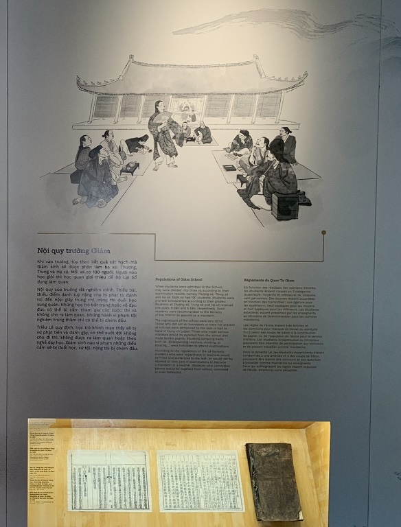 An exhibition wall with an ink illustration of students and a teacher in an old academy hall, text panels below and old books in a glass case