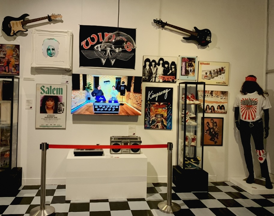A museum wall of 1980s music memorabilia: electric guitars, band posters, a boombox, sneakers in a glass case and a mannequin in a band T-shirt above a checkerboard floor