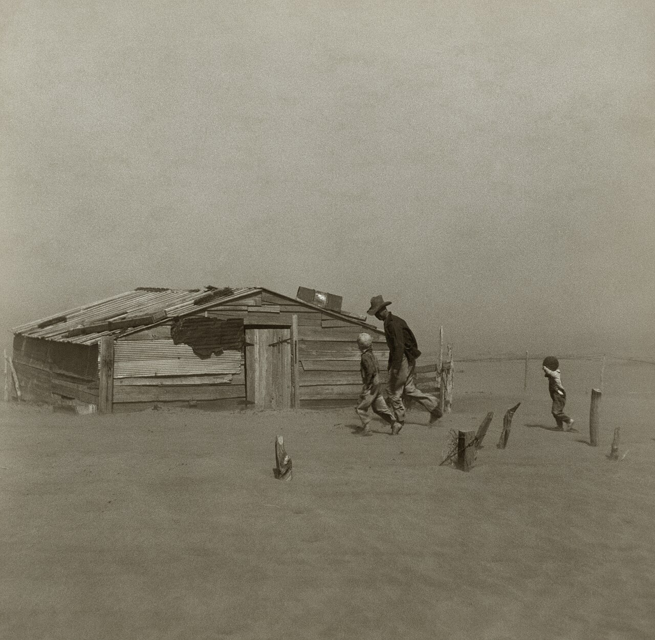 A father and two sons walking past a half-buried shed in a dust storm