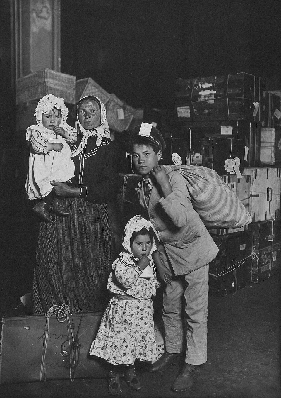 A mother holding a baby, a boy carrying a large bundle on his back and a little girl holding his hand, surrounded by luggage