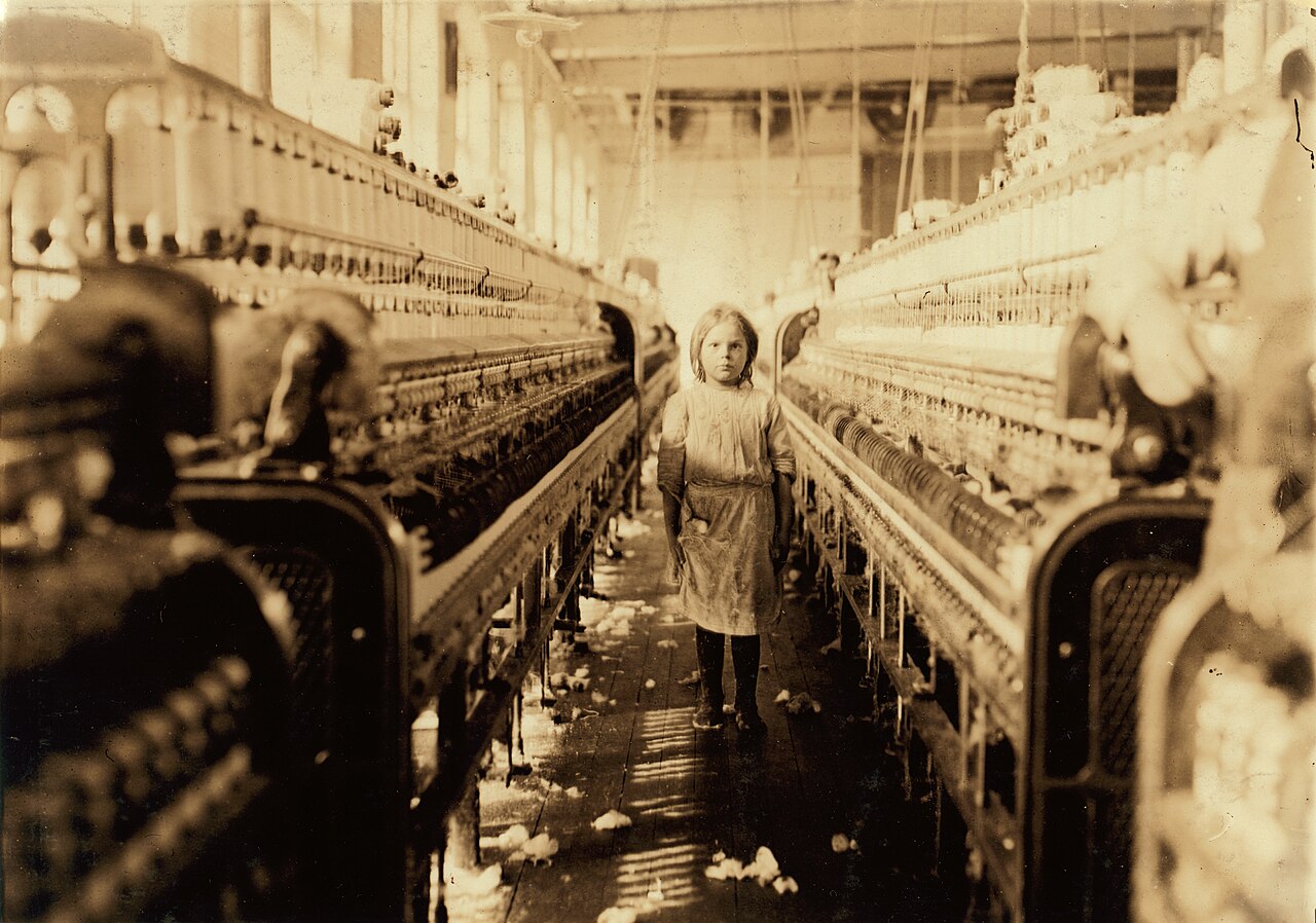 A small girl standing in the aisle between huge spinning machines in a cotton mill
